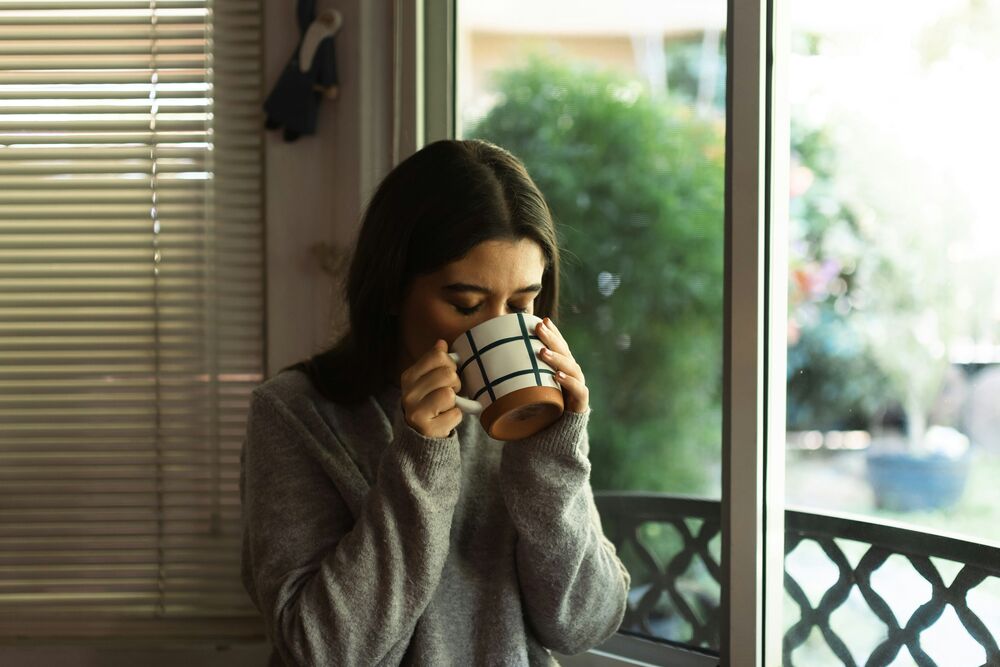 Femme buvant une tasse chaude près d'une fenêtre, moment de calme et de récupération