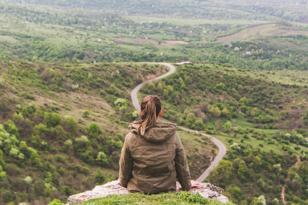 Femme assise sur une falaise, regard tourné vers une vallée verdoyante traversée d'une route sinueuse